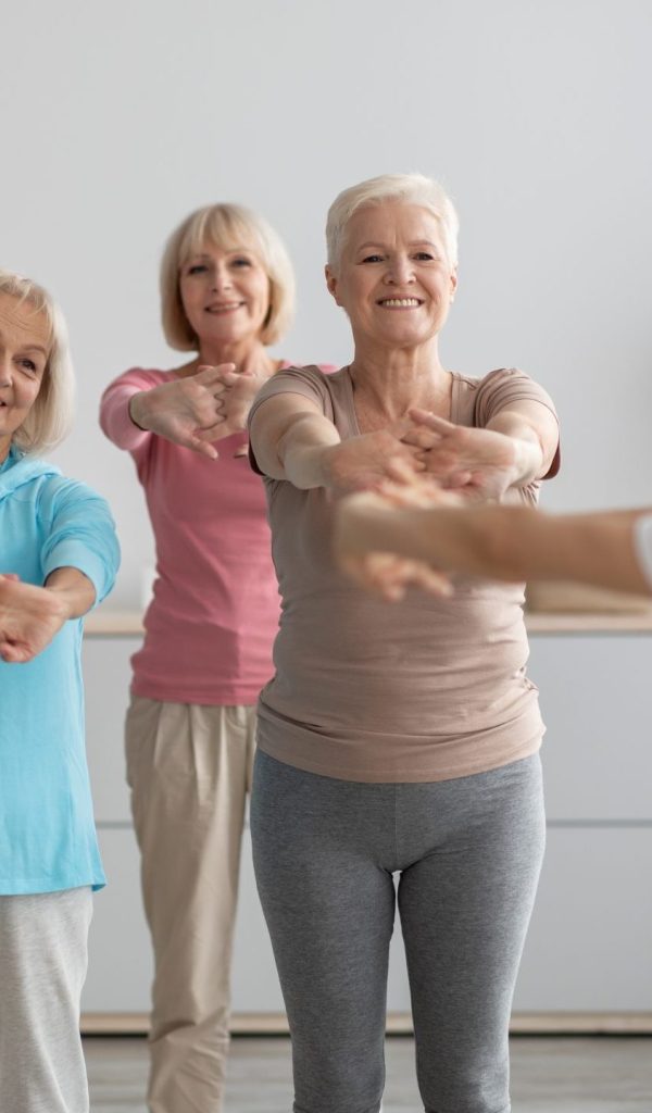 Sporty multiracial group of elderly people having fitness class at nursing home, training with female instructor, standing in a row, stretching hands and smiling, enjoying physical activity, panorama
